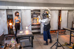 two glass students, one in protective gear, collaborate on a glassblowing piece