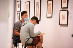 2 students sit and sketch in front of a row of artworks hanging in the risd museum