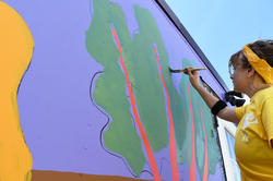 Graduate Student Simone Nemes stands on a ladder while painting stalks of kale on a shipping container at ECO City Farms