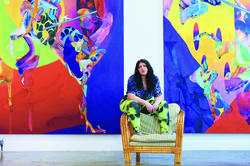the artists poses in front of two large, brilliantly colored paintings