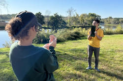 two students hold mirrors to reflect the sun next to the Blackstone River