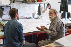 A graduate student and an educator discuss work over a busy desk space.