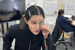 a student looks through a lens attached to several wires, in a classroom setting with a person at a desk in the background