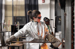 student wearing silver protective coat and protective glasses works with a boro rod inside a glass studio