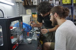 two people look closely as they work on a robotic object at a workstation equipped with other technological objects