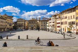 a city square in Florence, Italy, with people moving on foot or bike, under a blue sky with white clouds