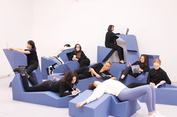 several students sit or lounge atop light blue, angular furniture inside an all-white room