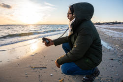 student dressed in a parka crouches down at the beach to record waves