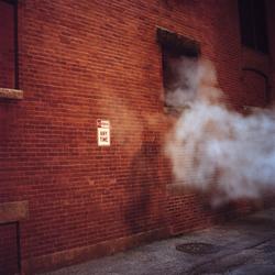 Student work by John Shen BFA 2019. Photograph of the side of a redbrick building in a steamy alleyway. 