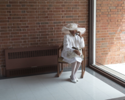 Student work by Nathan Miller MFA 2017. Photograph of a well-dressed woman talking on the phone while sitting in a chair in a sun-lit room.