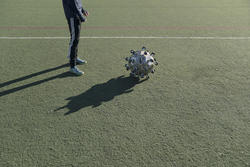 Student work by Sam Lavoie BFA 2018 ID/SC. A man standing on a soccer field in front of a metallic soccer ball. Each side of the ball has a small wheel protruding from it.