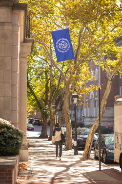 a blue flag with the RISD seal hangs from the front of Benson Hall, as a student walks on the sidewalk toward trees in the background