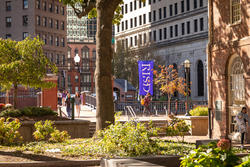 a blue flag with the letters RISD hangs in downtown Providence as students walk near green and gold foliage