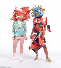two models wear colorfully patterned garments and sculptural masks designed by Desiree Scarborough