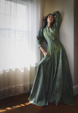 a model stands besides a window with light coming through a white curtain, wearing a long green dress designed by Emily Frisch