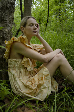 a model sits against a tree with forest greens behind her, wearing a pale yellow, patterned dress by Isabel Clarke