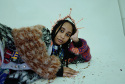 a model lies on the floor while wearing a fuzzy multicolored coat and a sculpted crown, all designed by Jackie Oh