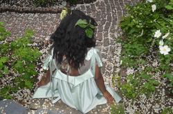 a model sits facing away from the camera, surrounded by greenery and white flowers, in a white dress by Joslynne Houlder