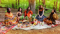 several models sit in a forest clearing, surrounding a picnic blanket, all wearing designs by Mina Serbetcioglu