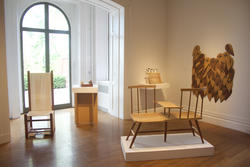 light-brown wooden chairs and other furniture sit in a gallery room illuminated by light coming through a window in the background