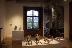 ceramic sculptures of fantastical creatures sit on a display surface at the center of a gallery room, dimly illuminated by light from a rear window