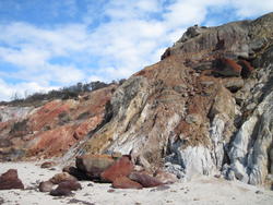 a cliff face of red, brown and gray rock running along a sandy beach, with slightly cloudy blue sky in the background