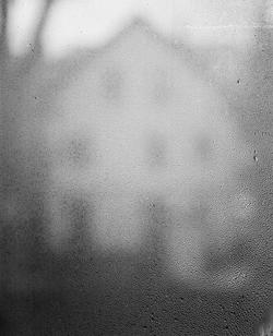 black and white photo of a white house with several windows, out of focus behind a glass pane wet with rainwater