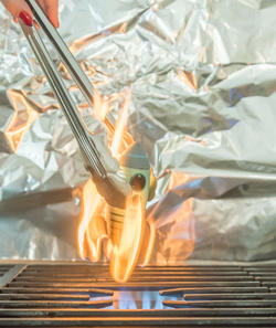 metal tongs hold a miniature rocket over a lit stove flame, fingertips with red nail polish visible against a silver foil background