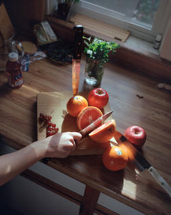 a knife held in someone's hand slices through a grapefruit on a cutting board, surrounded by other fruit, placed atop a wooden counter