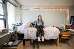 student sits on bed with floral print cover inside dorm room with white string lights on white wall