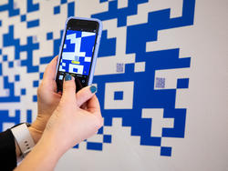 hands hold a smartphone up to a large QR code printed on a while wall, with yellow popup visible on phone to show its scanning the QR