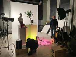 a student kneels on the floor in front of several arranged objects, in preparation for a photo shoot, with faculty and classmates looking on