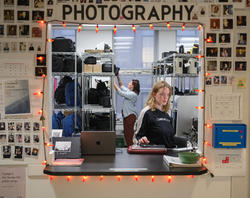 a student at an open window (surrounded by string lights and with the word "photography" written above) works at a desk as another student pulls equipment off a shelf in the background