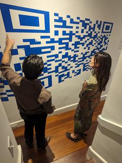 a student discusses a blue and white, large-scale QR code artwork to a gallery visitor