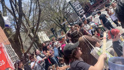 A crowd of people stand together outdoors while playing percussion instruments as part of a protest demonstration