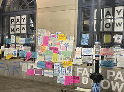 many colorful fliers hang on the outside walls of a building with the words "PAY NOW" displayed in a pair of windows