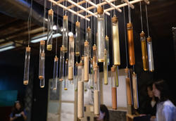 more than 20 test tubes filled with different colored materials hang suspended from a ceiling inside a dark room with students standing in the background