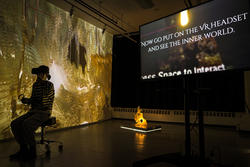 a student wearing a virtual reality headset sits in a chair in dark, gold-tinged light, with a nearby tv monitor reading "Now go put on the VR headset and see the inner world."