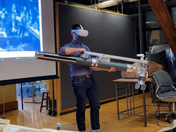 a student wearing virtual reality goggles holds a large robotic arm inside a dark studio space, with an active projector screen behind him