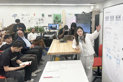 a student points at presentation images on a whiteboard as an audience looks on to the left