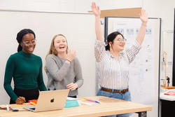 three students stand at a table laughing, with a student on the right holding their arms up high, looking at something outside the frame
