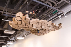 an overhead structure made of dozens of small brown blocks hangs overhead in a gallery space