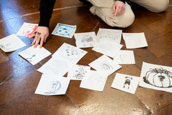 An array of drawings on white sheets of paper pile on the floor of a classroom