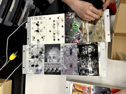 a hand manipulates a device featuring eight punk zine-style images, including one of Albert Einstein, with holes for audio cables prominent along the surface