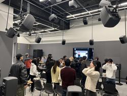 several students, many with their ears cupped, stand inside a studio space with many speakers hanging from the ceiling