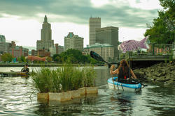 a structure sprouting greenery floats in a river as people row kayaks around it, with the downtown Providence skyline in the background