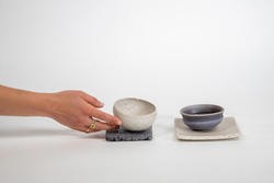 a hand reaches toward a white bowl on top of a gray coaster, sitting next to a gray bowl on a white coaster