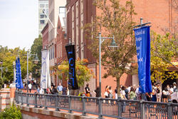 one white flag and one blue flag, bearing the letters R I S D, fly along a riverwalk as people gather on the outside of a brick building