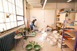 a student places a green ceramic pot near the wall of a studio filled with green and white ceramic vessels
