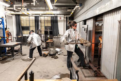 two students wearing silver protective gear work with equipment inside a glassblowing studio